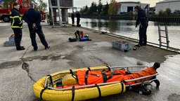 Am Rande eines Hafenbeckes stehen Feuerwehrmänner. Im Vordergrund liegt ein gelbes Schlauchboot mit einem Dummy darin. 