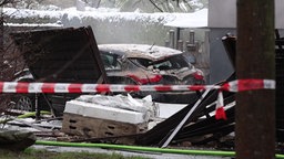 Ein zerstörtes Auto steht hinter einem Absperrband vor dem Haus