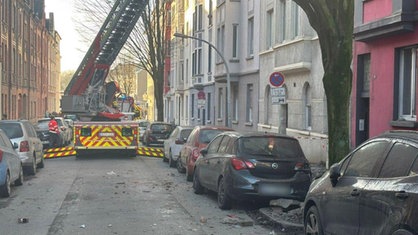 Eine Straße, auf die teile des Hauses gestürzt sind. Ein Feuerwehrwagen steht mit einer Leiter vor dem Haus, an den Straßenrändern parken Autos. 