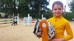 Jette steht mit ihrem Steckenpferd Milo vor dem Springparcours den Hobby Horsing Cups. Milo trägt eine goldene Schleife.