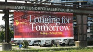 An der Jahrhunderthalle in Bochum hängt en großes Plakat für die Ruhrtriennale 2024 mit dem Titel "Longing for Tomorrow"