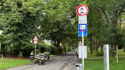 Polizisten stehen am Wegesrand in einer Straße, davor ein Schild, das Sperrzeiten für den motorisierten Verkehr hier anzeigt.