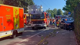 Rechts ein verunfallter PKW, auf dem Gehweg sind Kreidemarkierungen zu sehen. Daneben stehen Wagen des Rettungsdienstes und der Feuerwehr.