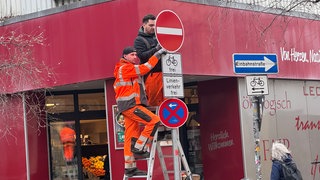 Zwei Arbeiter stehen auf einer Leiter und montieren von einem Mast ein Schilder ab