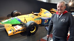 Reiner Ferling, Vorsitzender des Michael und Mick Schumacher Fan-Club Kerpen, steht zwischen Rennwagen, die Michael Schumacher gefahren hat