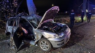 Ein weitgehend zerstörter grauer VW Polo, im Hintergrund Polizisten