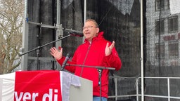 Ein Mann in roter Jacke auf einer Bühne hinter einem Rednerpult, das mit einer Verdi-Flagge umhüllt ist