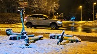 Mehrer E-Scooter liegen im Schnee, im Hintergrund fährt ein Auto.