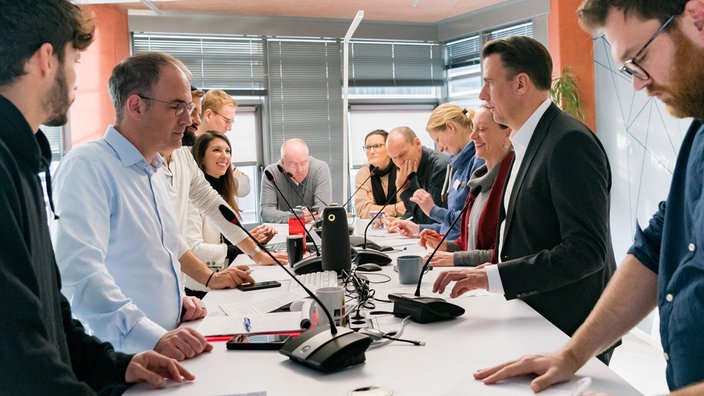 Frauen und Männer stehen an einem großen Konferenztisch mit Mikrofonen