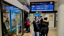 Menschen befinden sich innen im Bahnhof unter der Anzeigetafel.