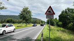 Man sieht eine Straße, an deren Seite ein dreieckiges Warn-Schild mit einem Biber Piktogramm steht. Darunter befindet sich die Aufschrift: "Biberwechsel 19-7h".