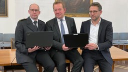 Drei Herren im Anzug sitzen auf einem Tisch und halten zwei Laptops zwischen sich.