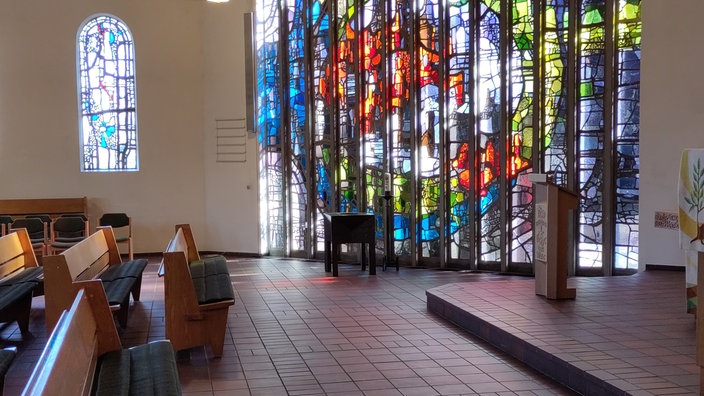 Innenraum einer Kirche mit einem großen Kirchenfenster, durch das die Sonne scheint. 