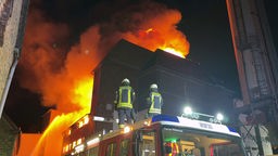 Feuerwehrleute stehen auf einem Feuerwehrwagen und schauen auf ein brennendes Gebäude.