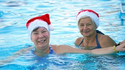 Zwei ältere Frauen sind in einem Schwimmbad. Sie tragen weihnachtliche Mützen.