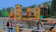 Ein großer Burg-ähnlicher Erlebnisspielplatz aus Holz ist umgeben von einem kleinen Wasserpark. Viele Besucher tummeln sich auf den Türmen des Spielplatzes und auf kleinen Floßen und Stegen.