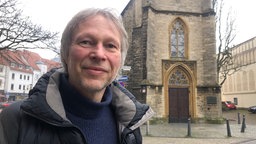 Ein Mann in Pullover und Jacke vor einem Kirchengebäude