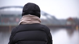 Eine Person sitzt mit dem Rücken in der Landschaft, im Hintergrund eine Brücke