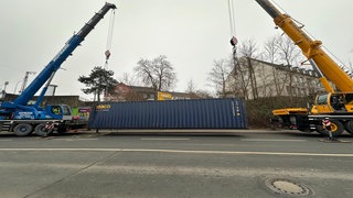 Ein Container wird von zwei Kränen hochgezogen.