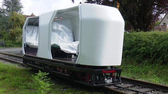 Ein Monocab - ein weißes, längliches Fahrzeug - fährt auf einer Schiene einer Zugstrecke.