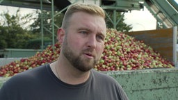 Ein junger Mann mit gestutztem dunkelblondem Bart steht vor einem Anhänger voller Äpfel