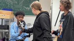 Eine Schülerin reicht ihrem Mitschüler eine Fahrradkurbel samt Pedal. Sie befinden sich im Klassenzimmer.