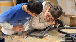 Zwei Schüler lehnen sich über eine Werkbank, auf der Fahrrad-Schläuche liegen. Der rechte hält einen Schlauch an sein Ohr.