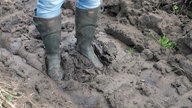 Auf dem Bild sind Gummiestiefel zu sehen, die im matschigen Boden des Feldes eingesunken sind