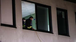 Ein Mann mit blauer-neongelber Uniform schaut aus einem Fenster. Er hält ein Kabel in der Hand, das an der Hauswand nach unten hängt.