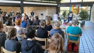 Menschen sitzen bei der Trauerfeier von Malte in einem Saal mit Stühlen. 