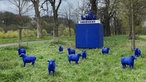 Zu sehen sind blaue Schafsstatuen, welche auf einer Wiese stehen, hinter ihnen steht ein blauer Quader auf welchem vier Schafe einen Streitwagen ziehen, wo der Fahrer ein Peace-Zeichen emporhält.