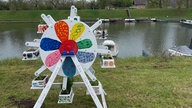 Zu sehen ist ein Miniaturriesenrad, welches bunt bemalt ist. Es steht auf einer Wiese vor einem See.