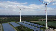 Zwei Windräder stehen auf einer freien Fläche, darunter Solaranlagen.