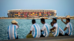 Fans in argentinischen Trikots sitzen am Wasser. Im Hintergrund das Stadium 974