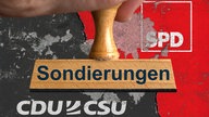 Ein symbolischer Holzstempel mit der Aufschrift "Sondierungen", gehalten von einer Hand, vor einer Wand mit den Logos von CDU / CSU (Union) und SPD auf den abgeplatzten Farben Schwarz und Rot sowie einem dicken Riss.