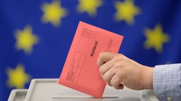 Symbolbild: Vor einer EU-Fahne steht eine Wahlurne, in die eine Person einen Wahlbrief einwirft.