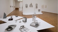 Fluxus and Beyond: Werk von Ursula Burghard in der Ausstellung. Silberne Alltagsobjekte, wie Schuhe, Taschen, etc. auf Tischen und Wänden. 