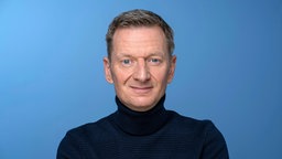 Der Schauspieler Michael Kessler in schwarzem Pulli vor blauem Hintergrund.