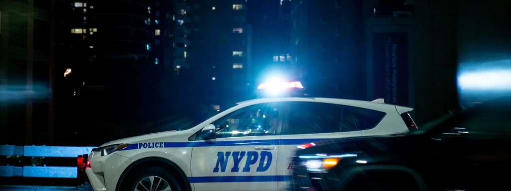 Ein Polizeiauto fährt mit Blaulicht durch die Straßen New Yorks, auf dem Auto steht in blauer Schrift "Police" und "NYPD".