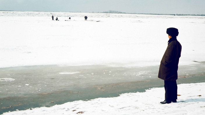 Ein Mann mit Pelzmütze und Winterkleidung steht am Ufer des zugefrorenen Amur nahe Chabarowsk und schaut in Richtung einer Gruppe Eisfischer.