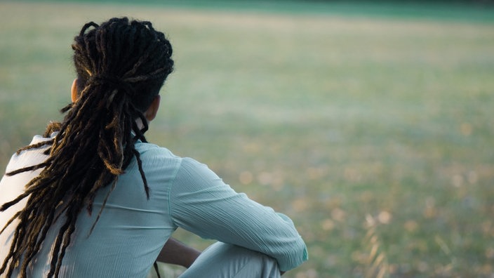 Oberkörper eines Mannes mit Dreadlocks, der auf einer Wiese sitzend von hinten