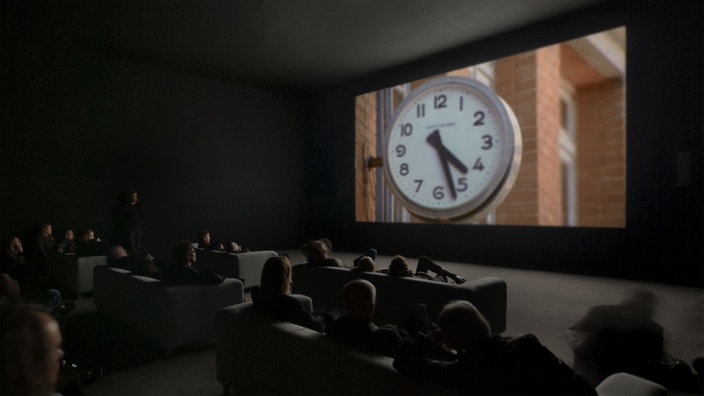 Filminstallation "The Clock" im Kunstmuseum Stuttgart: Zuschauer in einem Vorführraum blicken auf die Projektion einer Uhr.