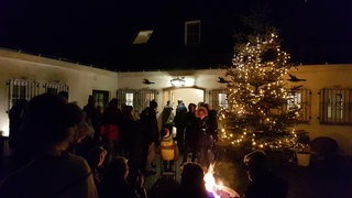 Lebendiger Adventskalender in Hülsenbusch: Menschen stehen versammelt um einen geschmückten Weihnachtsbaum und eine Feuerstelle.