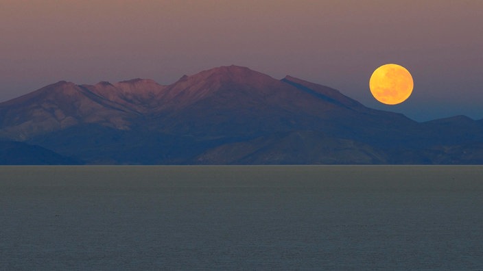 Vollmond im Morgengrauen, der hinter sich hinter einem Berg am Meer absenkt.