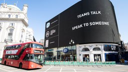 Ein roter Bus vor einer großen Leuchtschrift am Piccadilly Circus mit der Aufschrift: "Teams Talk - Speak to someone".