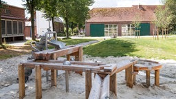 Außenansicht von Gebäuden, davor eine hüfthohe Wasser- / Kugelbahn aus Holz auf einer Sandfläche.