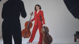 Cellistin Anastasia Kobekina steht in einem eleganten, roten Anzug mit ihren Instrumenten vor einer weißen Studioleinwand.