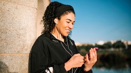 Lächelnde Frau in sportlicher Kleidung, die mit einem Smartphone Musik hört, während sie an einer steinernen Säule lehnt.