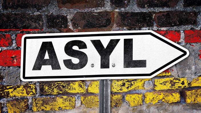 Ein Schild mit der Aufschrift "Asyl" vor einer schwarz/rot/gold bemalten Mauer