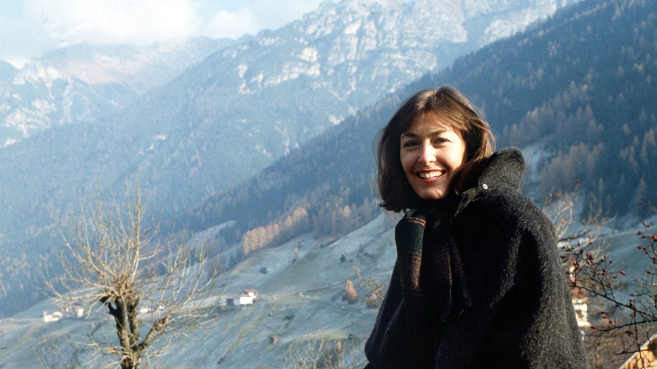 Thekla Carola Wied sitzt auf einem Stein, im Hintergrund sieht man ein Dorf in den Bergen